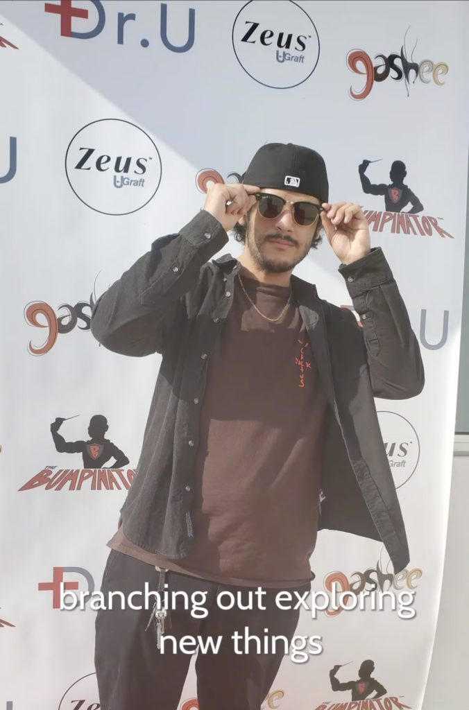 Young adult Alejandro wearing a black cap and sunglasses while standing in front of a Dr. U branded event backdrop.