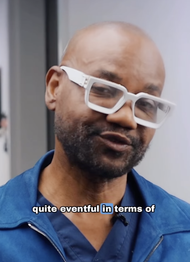 Dr. Sanusi Umar wearing white glasses and blue clothing while speaking on camera with subtitle text across the lower portion of the frame.