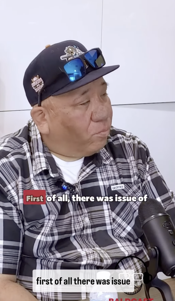 A man wearing a black cap, blue sunglasses, and a plaid shirt speaks into a tabletop microphone.