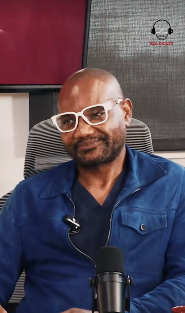 Dr. Sanusi Umar seated in a blue jacket and white glasses with a microphone in front of him.