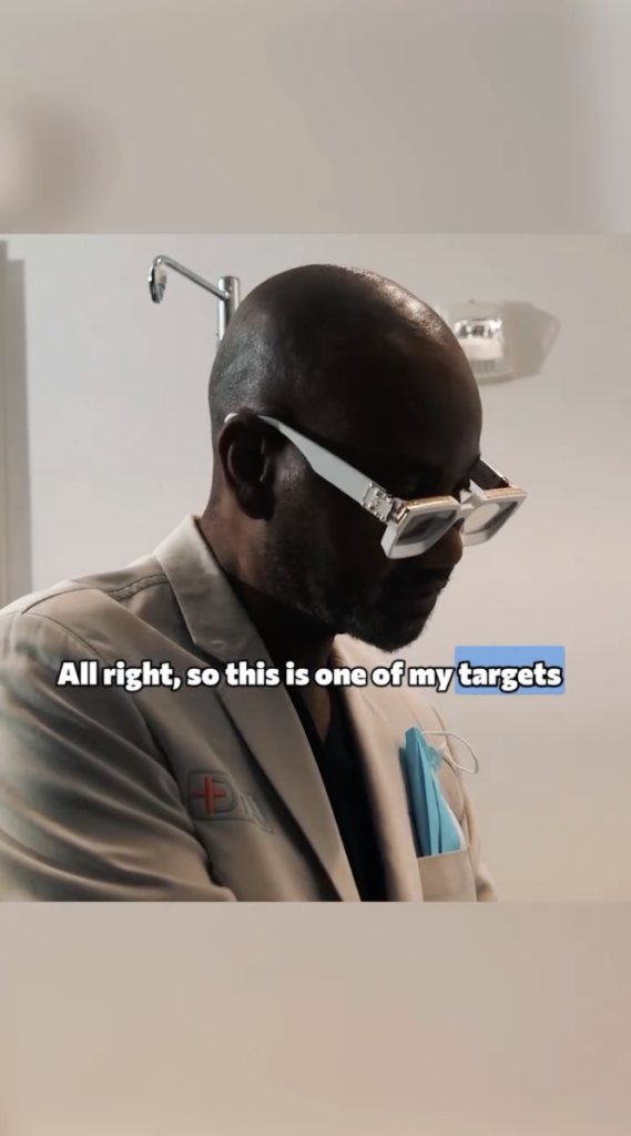 Dr. Sanusi Umar wearing white glasses and a light-colored medical jacket in a clinical room.