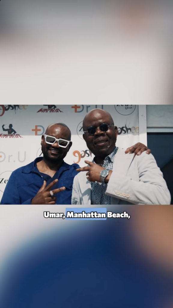 Dr. Sanusi Umar and Festus standing together, smiling and making peace signs in front of a Dr. U and Bumpinator backdrop.