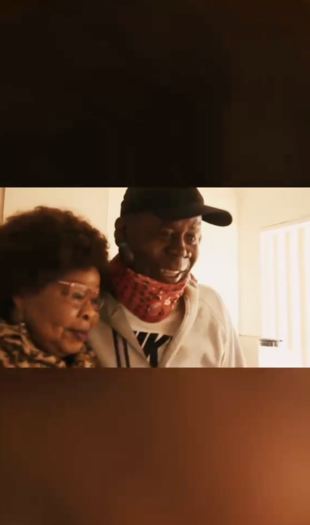 Festus wearing a black cap and light jacket standing closely beside a woman indoors.