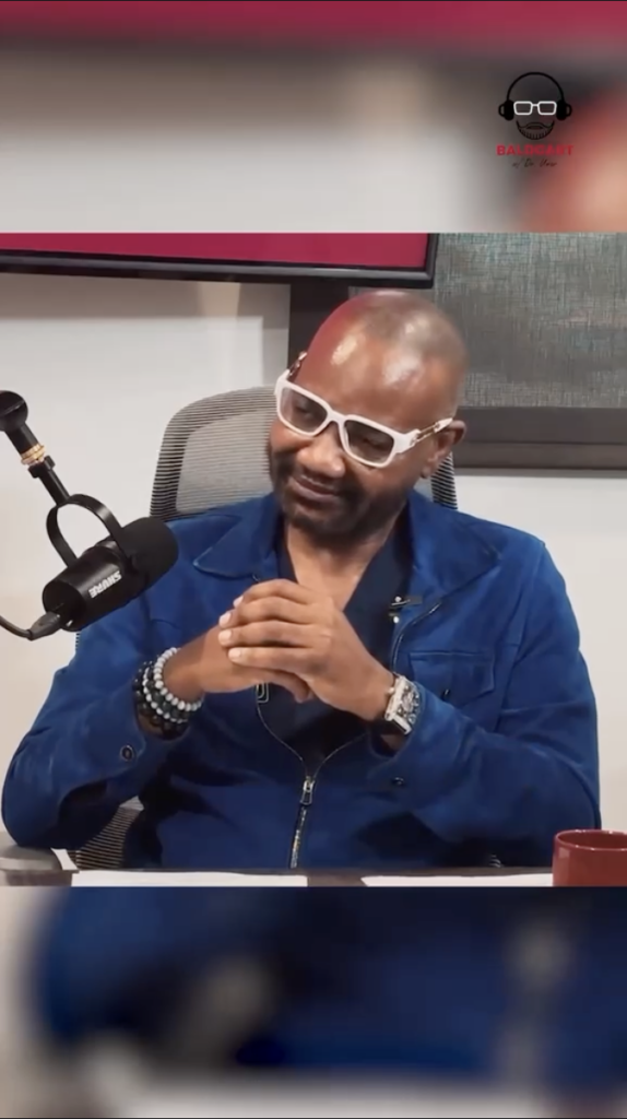 Dr. Sanusi Umar sits at a podcast table wearing white glasses and a blue jacket with a microphone beside him.